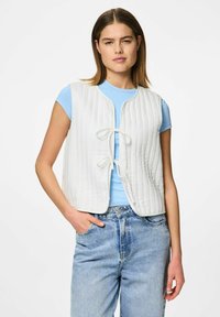 Vit vadderad väst med en strukturerad, kviltad design, med en frontknäppning och rund halsringning, bärs över en ljusblå figurnära skjorta och jeans.