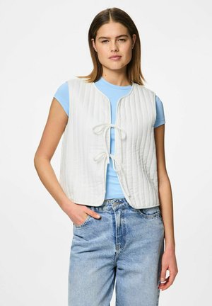 Hvid polstret vest med et tekstureret, quiltet design, der har en foranbånd og en rund halsudskæring, båret over en lyseblå tætsiddende skjorte og denimjeans.