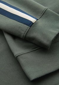 Manga de sudadera verde oscuro con rayas en contraste en azul marino y blanco. La textura de puño acanalado añade detalle. El ángulo en primer plano enfatiza el diseño.