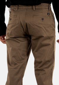 Brown casual pants with buttoned back pockets worn by a person in a black long-sleeve top, showing the waist and upper leg area.