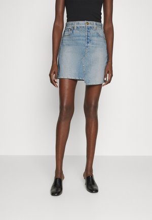 Femme portant un haut noir sans manches, une mini-jupe en denim asymétrique bleu clair et des chaussures noires sans lacets, debout devant un fond blanc.