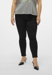Crne skinny traperice s glatkom teksturom i srednjim strukom, uparene s labavom sivom majicom i crnim cipelama s oštrim vrhom.