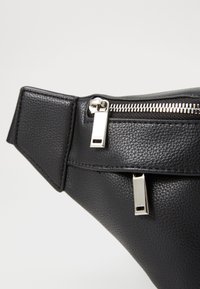 Zwarte leren heuptas met een gestructureerde afwerking; voorzien van zilverkleurige metalen ritshardware en een verstelbare riem.