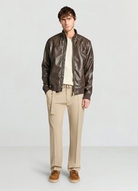 Veste aviateur en cuir marron, t-shirt beige, pantalon beige avec un détail latéral, et chaussures marron. Le mannequin pose devant un fond neutre.