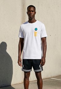 Mladi moški stoji ob betonskem zidu, oblečen v belo majico s košarkaškim potiskom in črne športne kratke hlače z belim robom.