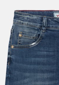 Jeans en denim bleu foncé avec une texture lisse, des accessoires dorés et un design classique à cinq poches. Détail rouge à l'intérieur de la taille.