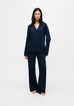 Femme debout pieds nus portant un haut bleu foncé à manches longues boutonné et un pantalon large assorti sur un fond clair uni.