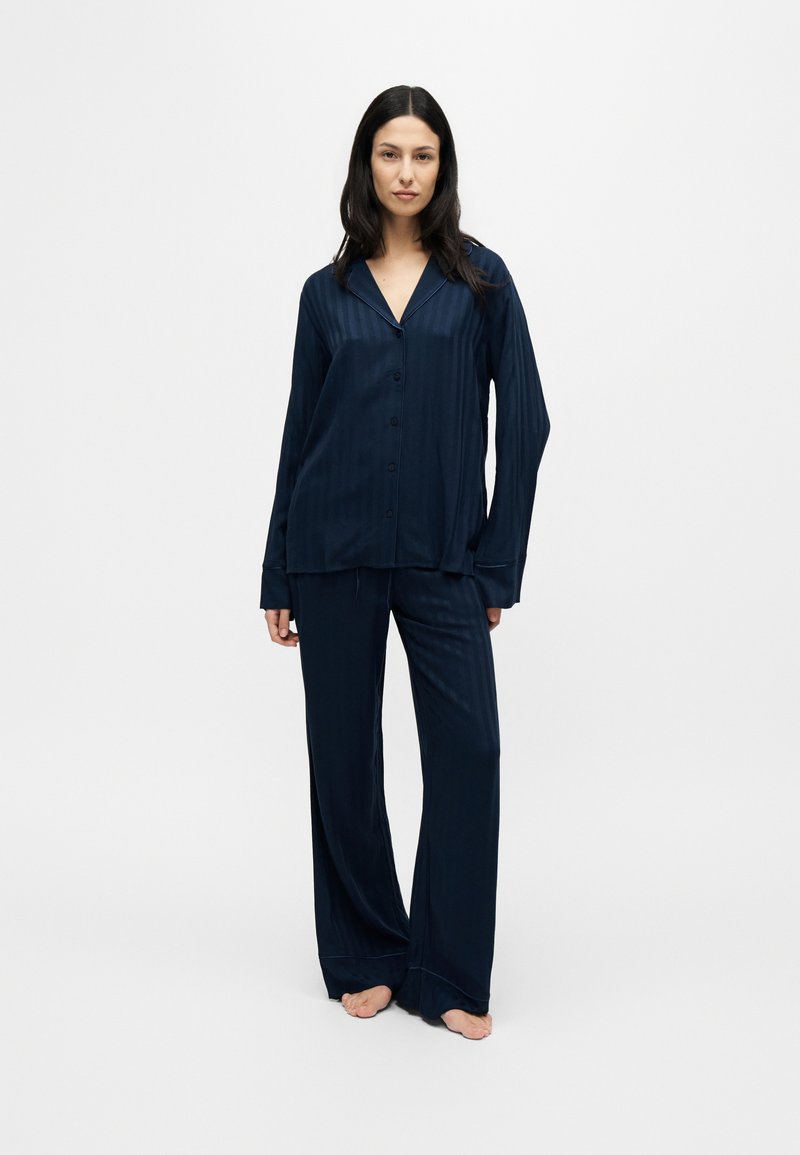 Femme debout pieds nus portant un haut bleu foncé à manches longues boutonné et un pantalon large assorti sur un fond clair uni.
