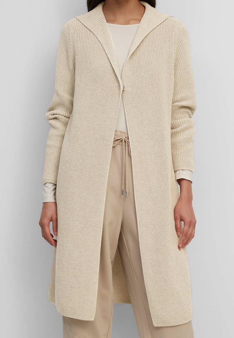 Cardigan beige a maglia con un ampio cappuccio, apertura frontale e maniche lunghe. Il tessuto testurizzato conferisce un aspetto accogliente; abbinato a pantaloni beige.
