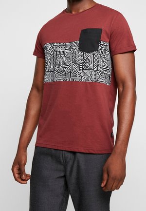 Bordeauxrood T-shirt met een geometrische zwart-wit print over de borst, voorzien van een zwart zakje aan de linkerkant. Korte mouwen.