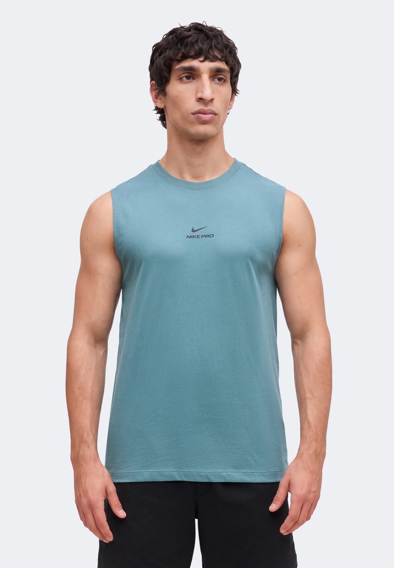Άνδρας που φοράει μπλε αμάνικη μπλούζα Nike Pro και μαύρα σορτς, στέκεται μπροστά σε απλό φωτεινό φόντο, κοιτώντας μπροστά.