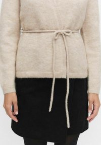 Jasno beżowy sweter z dzianiny z wiązaniem w talii i ryflowaną teksturą. Połączony z czarną spódnicą z sztruksu, podkreślającą kontrastujące materiały.