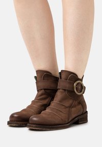Botins de couro castanho até ao tornozelo com ponta arredondada, design solto, uma faixa larga com uma fivela circular metálica e um salto baixo em camadas.