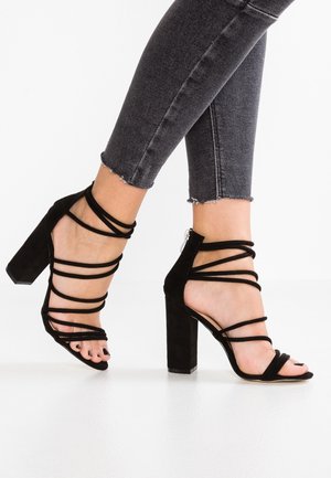 Fødder i sorte rem-sandaler med høj hæl, stylet med slidte mørkegrå skinny jeans på hvid baggrund.