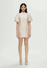 Nő világos bézs, rövid, puffos ujjú ruhában, texturált virágmintával, karjaival az oldalán áll egy sima háttér előtt.