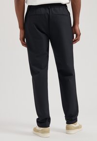 Zwarte, taps toelopende broek met een gladde textuur en elastische tailleband. Voorzien van twee achterzakken. Gestyled met lichtgekleurde sneakers.