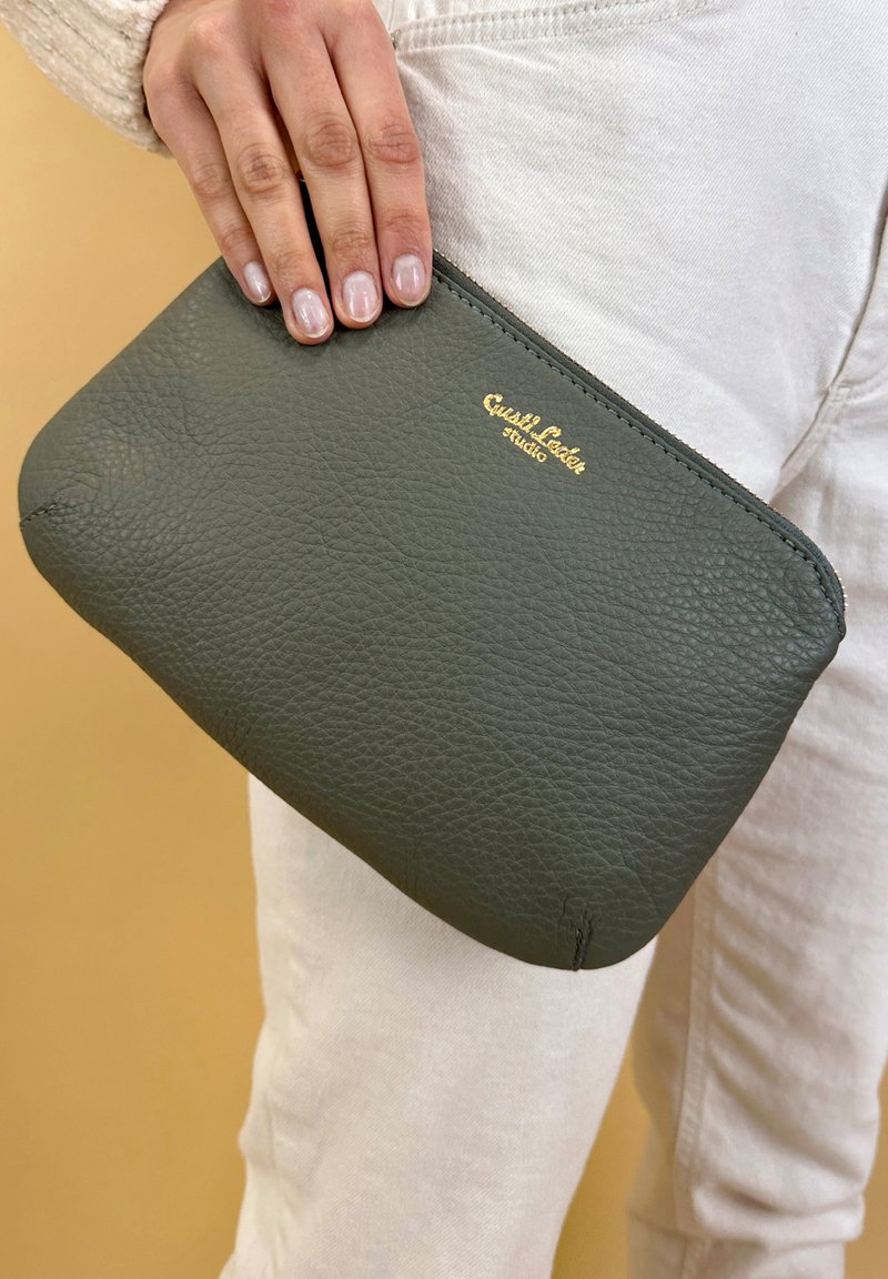 Groene leren clutch met een textuur, ritssluiting en goudkleurig reliëflogo. Vastgehouden in een hand tegen een neutrale achtergrond.