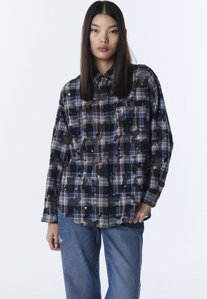 Camisa de cuadros negros y azules con un patrón de cuadros grandes, diseño abotonado, dos bolsillos en el pecho y un ajuste relajado. La tela parece texturizada.