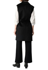 Schwarzer ärmelloser Mantel aus strukturiertem Stoff, mit tailliertem Gürtel und Kragen, kombiniert mit einem weißen Langarmhemd und schwarzen Hosen.