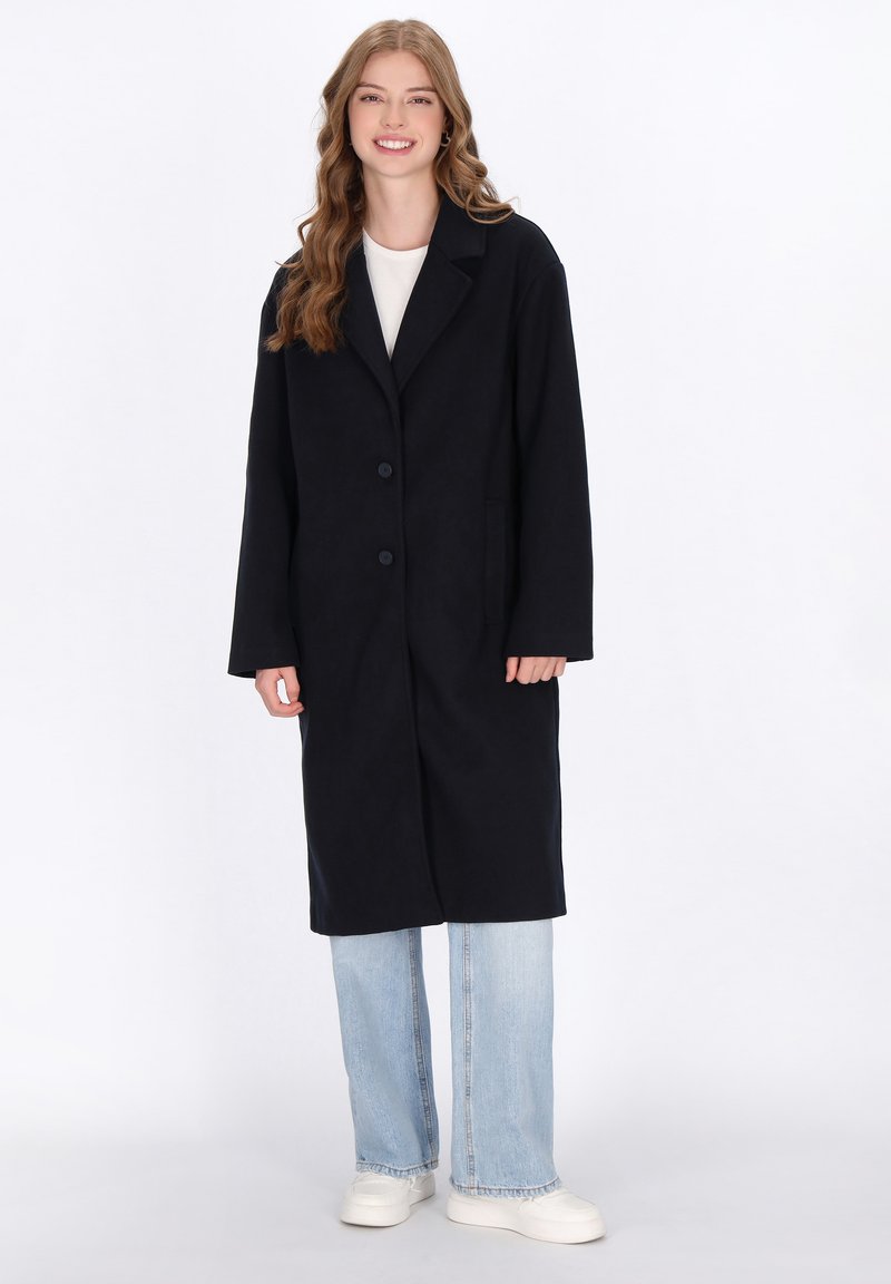 Cappotto lungo nero con colletto a punte, due tasche frontali e chiusura a bottoni, abbinato a jeans azzurri chiari e sneaker bianche.