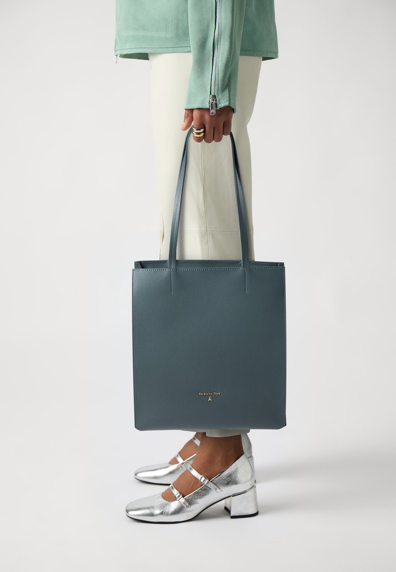 Square green leather tote bag with dual handles and gold logo; paired with silver Mary Jane shoes featuring block heels and ankle straps.