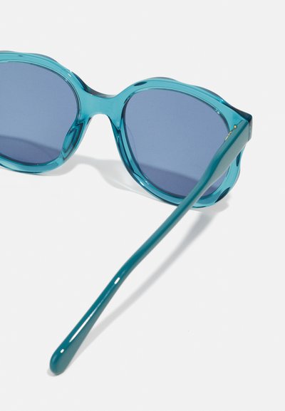 Occhiali da sole in plastica turchese con lenti rotonde di grandi dimensioni. Presentano una montatura spessa e ondulata e aste sottili e dritte. Le lenti sono di colore blu scuro.