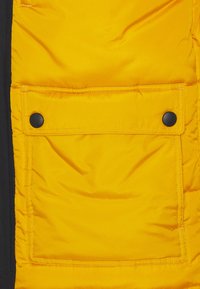 Gul polstret jakkeafsnit med en stor lomme med sorte trykknapper, der står i kontrast til en mørk stofbaggrund.