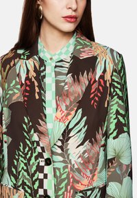 Blazer mit floralem Muster in Dunkelbraun, mit Grüntönen, Rot und Beige. Darunter befindet sich eine grün-karierte, knöpfbare Bluse. Glatte Textur.
