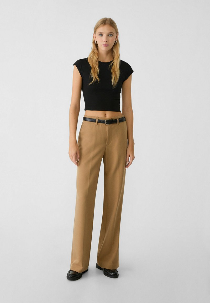 Donna in piedi con un top corto nero, pantaloni beige a gamba larga con una cintura nera, scarpe nere e orecchini a cerchio dorati su uno sfondo semplice.