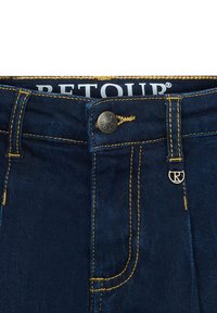 Jeans in denim blu scuro con cuciture gialle, un bottone in metallo, passanti per la cintura e un piccolo logo rotondo in metallo vicino alla vita.