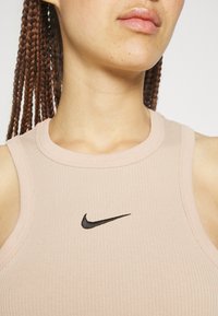 Gros plan d'une personne aux cheveux tressés portant un débardeur côtelé beige clair avec un petit logo noir Nike swoosh sur la poitrine.
