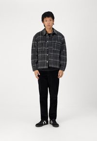 Grijze plaid knoopjacket met een ontspannen pasvorm, gedragen over een zwart shirt, gecombineerd met zwarte corduroy broeken en zwarte sneakers.