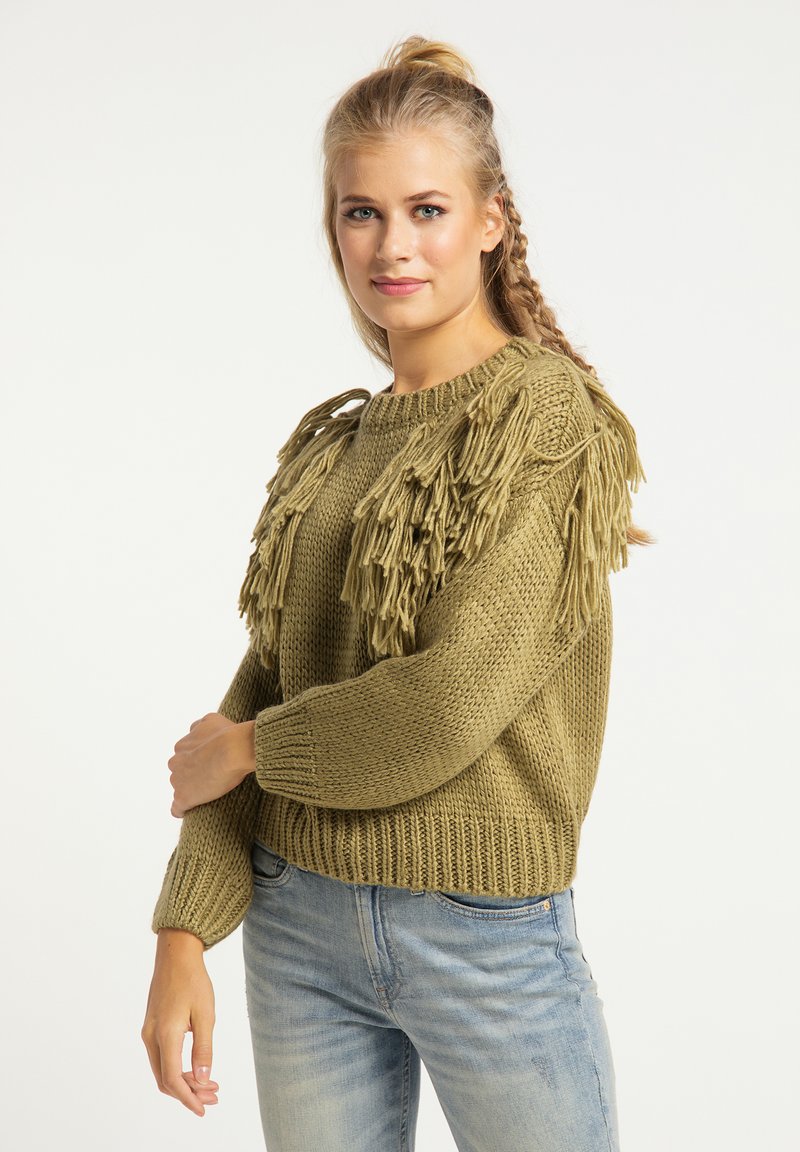Olijfgroene gebreide trui met franjes op de schouders, ronde halslijn en geribde manchetten. Gecombineerd met lichtblauwe jeans.