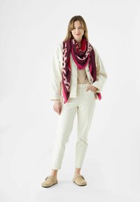 Beige spijkerjack over een lichtbeige top, gecombineerd met ivoorkleurige broeken en beige klompen. Een meerkleurige bordeauxrode sjaal voegt contrast en textuur toe.