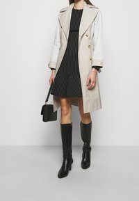 Béžový trenčkot se zlatými knoflíky přes černé pruhované šaty, doplněný nad kolena sahajícími černými koženými botami a malou černou kabelkou.
