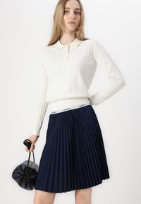 Cream ribbed long-sleeve polo shirt with three buttons, paired with a pleated navy skirt. Holding a black drawstring bag.