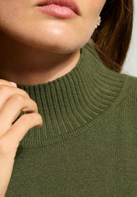 Nahaufnahme einer Person, die den gerippten Kragen eines grünen Strickpullovers berührt, mit sichtbaren Lippen, Kinn und einem Teil eines Ohrs mit einem Ohrring.
