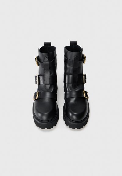 Botas de cuero negras hasta el tobillo con tres correas ajustables, hebillas doradas y una suela de goma gruesa. Acabado liso con punta redondeada.