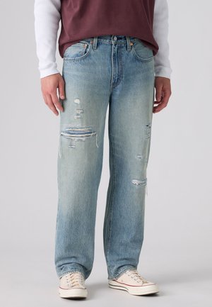 Homme portant un jean droit déchiré bleu clair, des baskets blanches, un haut bordeaux et une chemise blanche à manches longues, debout devant un fond uni.
