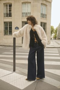 Purūs smėlinis paltas, juodas raukšlėtas viršus, plačios tamsios džinsinės kelnės ir smėliniai batai. Stovi greta perėjos su vertikaliu stulpeliu.