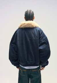 Marinblå överdimensionerad jacka med en fuskpälskrage, mjuk textur, vida ärmar och ribbad nederkant; matchad med mörka jeansbyxor med orange sömmar.