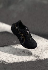 Black athletic shoe with mesh upper and metallic gold accents, featuring a breathable design and cushioned sole, suspended above a grey surface.