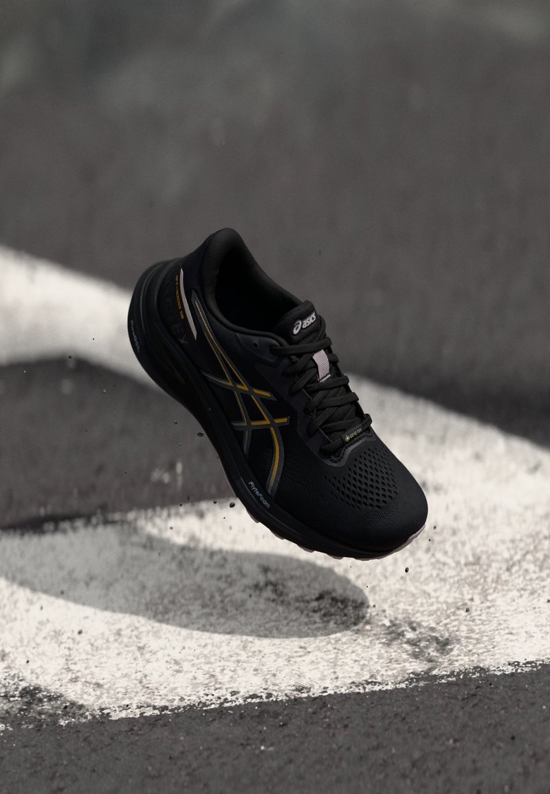Black athletic shoe with mesh upper and metallic gold accents, featuring a breathable design and cushioned sole, suspended above a grey surface.