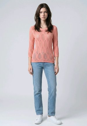 Mujer joven con cabello largo y castaño, que lleva un suéter de punto rosa, jeans azul claro y zapatillas blancas, posando frente a un fondo gris liso.