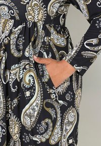 Musta mekko, jossa on beige, valkoinen ja vaaleansininen paisley-kuvio; teksturoitu kangas, pitkät hihat ja sivupocket-yksityiskohta.
