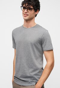 Νεαρός άνδρας με μαύρα ορθογώνια γυαλιά και απλό γκρι κοντομάνικο μπλουζάκι, στέκεται με τα χέρια στις τσέπες, χαμογελώντας ελαφρώς.