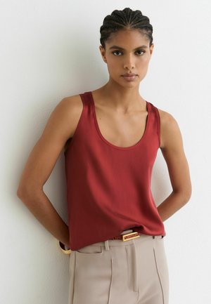 Kobieta z zaplecionymi włosami, ubrana w czerwoną, bezrękawnikową bluzkę i beżowe spodnie, stoi oparta o białą ścianę z rękami za plecami.