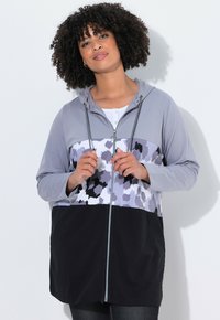 Felpa leggera con zip di colore grigio, caratterizzata da un design a blocchi di colore con un motivo floreale in tonalità di grigio e nero. Completata da un cappuccio con cordino.