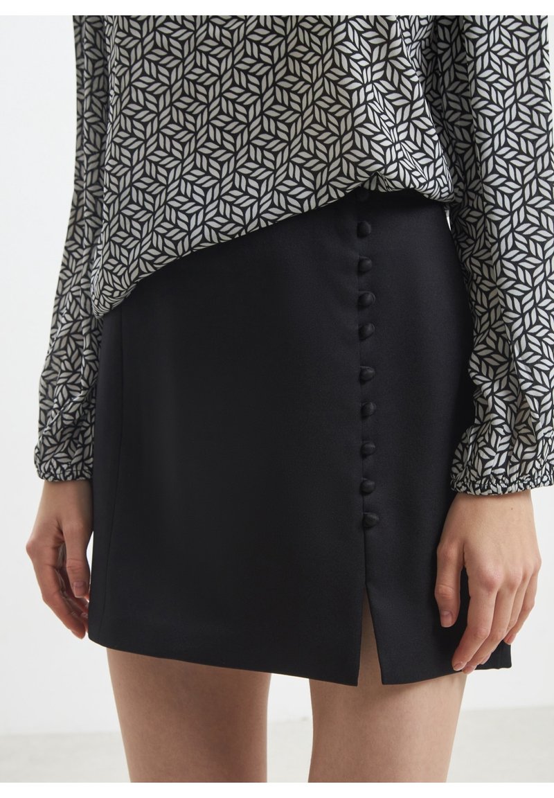 Jupe noire avec boutons décoratifs à l'avant et fente latérale, portée avec un chemisier à manches longues à motifs, mains détendues le long du corps.