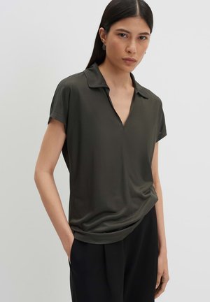 Femme aux cheveux lisses et foncés portant un haut vert foncé à manches courtes avec col et un pantalon noir, debout avec une main dans la poche devant un fond uni.
